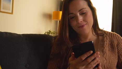 Laughing-Woman-Sitting-On-Sofa-At-Home-Using-Mobile-Phone-To-Check-Social-Media-Message-And-Scrolling-Online-5